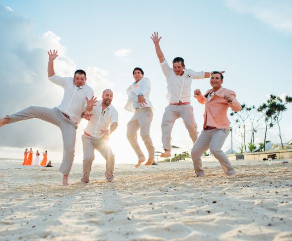 Five men in light-colored attire joyfully jump on a sandy beach at sunset. In the background, people in orange dresses, trees, and a cloudy sky are visible.