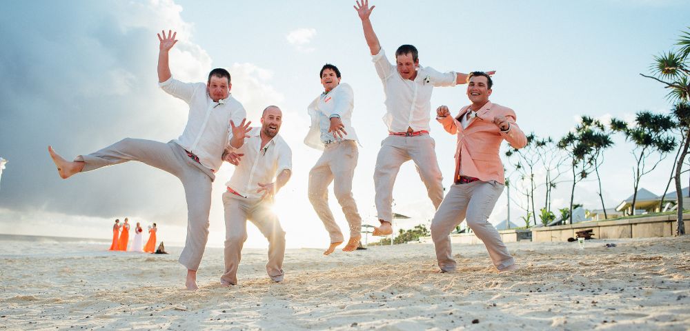Five men in light-colored attire joyfully jump on a sandy beach at sunset. In the background, people in orange dresses, trees, and a cloudy sky are visible.