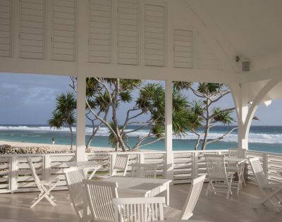 Bright beach pavilion with white furniture overlooking a sandy shore and turquoise ocean. Palm trees sway gently under a cloudy sky, evoking a serene atmosphere.