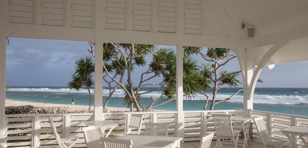 Bright beach pavilion with white furniture overlooking a sandy shore and turquoise ocean. Palm trees sway gently under a cloudy sky, evoking a serene atmosphere.
