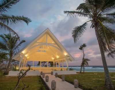 A white chapel with a peaked roof is illuminated at dusk, surrounded by palm trees with a pastel sky and ocean waves in the background. Tranquil atmosphere.