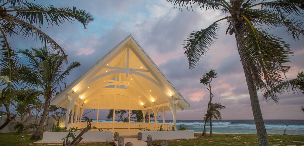 A white chapel with a peaked roof is illuminated at dusk, surrounded by palm trees with a pastel sky and ocean waves in the background. Tranquil atmosphere.