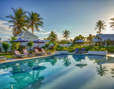 Tropical resort pool with clear water reflecting palm trees. Sun loungers with purple umbrellas line the pool, surrounded by lush greenery and cottages.
