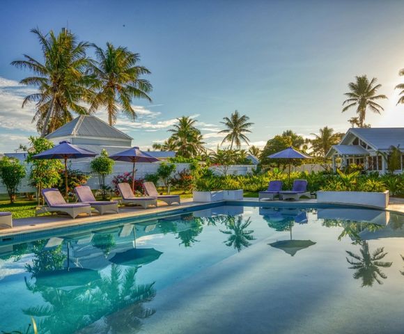 Tropical resort pool with clear water reflecting palm trees. Sun loungers with purple umbrellas line the pool, surrounded by lush greenery and cottages.