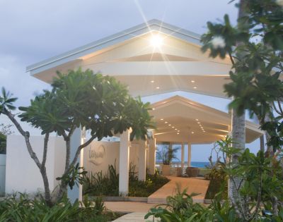 Tropical entrance with a lit canopy pathway, surrounded by lush greenery and tall trees. Ocean view in the background under a soft, cloudy sky.