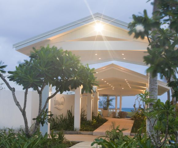 Tropical entrance with a lit canopy pathway, surrounded by lush greenery and tall trees. Ocean view in the background under a soft, cloudy sky.