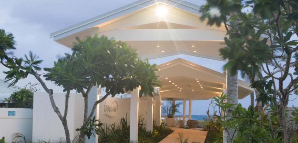 Tropical entrance with a lit canopy pathway, surrounded by lush greenery and tall trees. Ocean view in the background under a soft, cloudy sky.