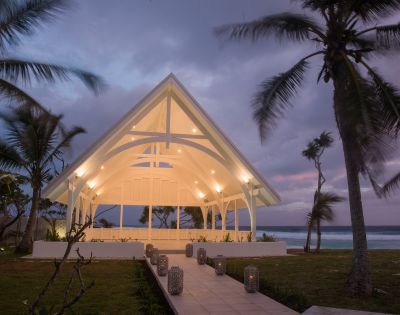 A modern, open-air chapel with a glowing, arched roof stands by the ocean at sunset. Surrounded by swaying palm trees, it creates a serene and peaceful ambiance.
