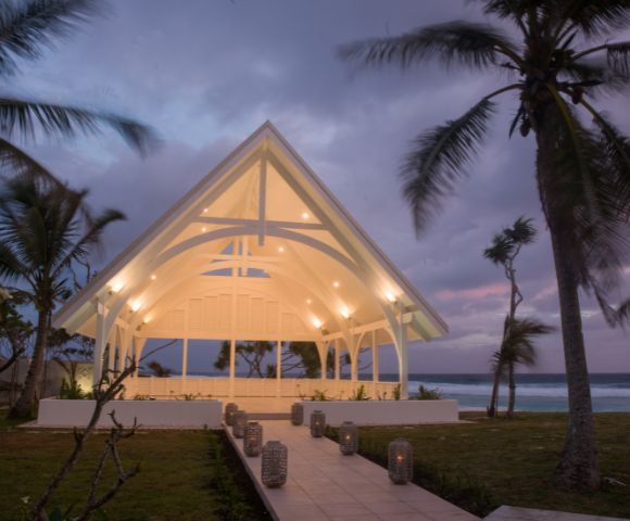 A modern, open-air chapel with a glowing, arched roof stands by the ocean at sunset. Surrounded by swaying palm trees, it creates a serene and peaceful ambiance.
