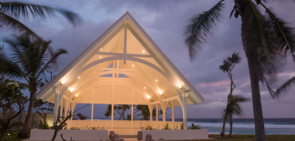 A modern, open-air chapel with a glowing, arched roof stands by the ocean at sunset. Surrounded by swaying palm trees, it creates a serene and peaceful ambiance.