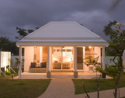 Cozy white cottage at dusk, warmly lit interior visible through large windows. Surrounded by lush greenery and palm trees under a cloudy sky.