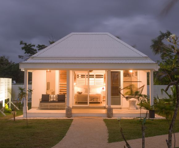 Cozy white cottage at dusk, warmly lit interior visible through large windows. Surrounded by lush greenery and palm trees under a cloudy sky.