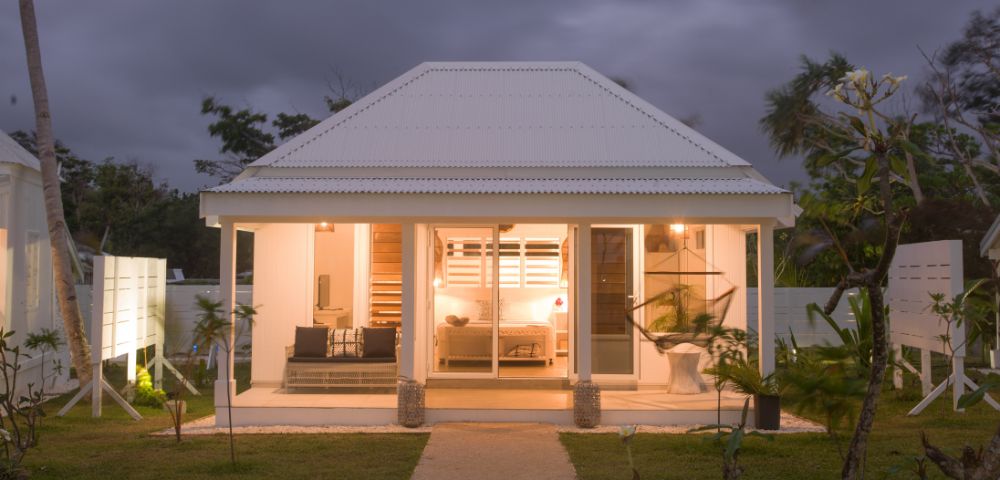 Cozy white cottage at dusk, warmly lit interior visible through large windows. Surrounded by lush greenery and palm trees under a cloudy sky.