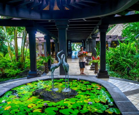 Serene tropical garden with lush greenery, a stone pond with lily pads and bird sculptures, and a person carrying fruit. A tranquil, welcoming atmosphere.