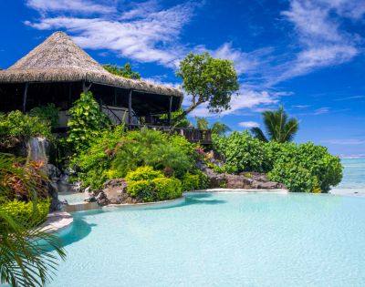 Tropical paradise scene featuring a thatched-roof hut surrounded by lush greenery, a cascading waterfall, and a serene turquoise pool under a bright blue sky.