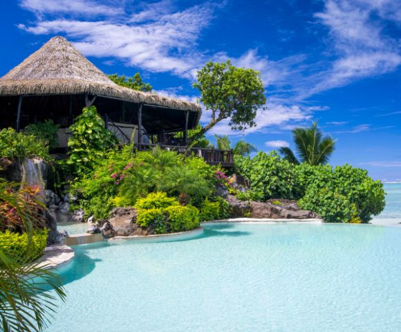 Tropical paradise scene featuring a thatched-roof hut surrounded by lush greenery, a cascading waterfall, and a serene turquoise pool under a bright blue sky.