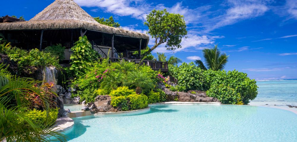 Tropical paradise scene featuring a thatched-roof hut surrounded by lush greenery, a cascading waterfall, and a serene turquoise pool under a bright blue sky.