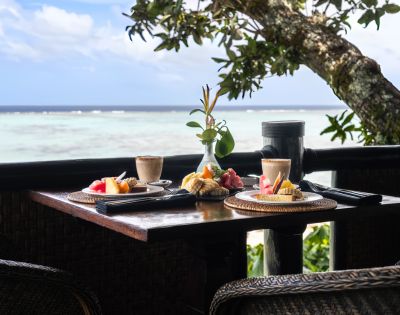 Breakfast table by a serene ocean view, featuring two plates of fresh fruit and pastries, lattes, and a small vase with greenery. Peaceful and inviting.