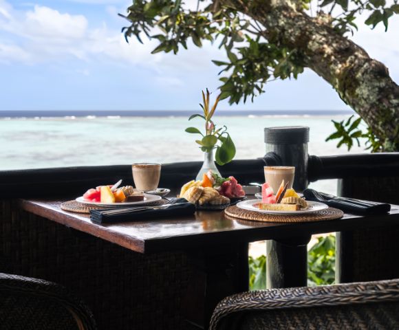 Breakfast table by a serene ocean view, featuring two plates of fresh fruit and pastries, lattes, and a small vase with greenery. Peaceful and inviting.