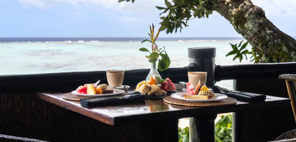 Breakfast table by a serene ocean view, featuring two plates of fresh fruit and pastries, lattes, and a small vase with greenery. Peaceful and inviting.