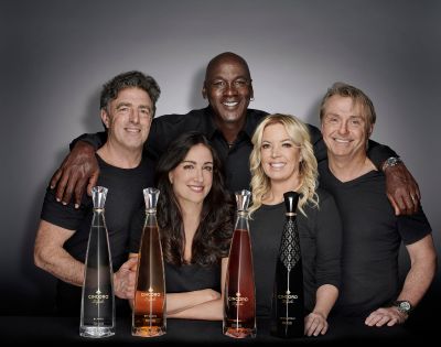 A diverse group of five people smiling warmly, posed behind four tall, elegant bottles of Cincoro Tequila. The setting is professional and inviting.