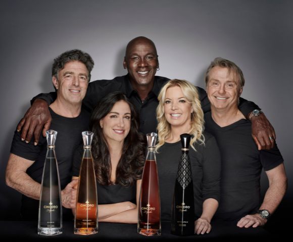 A diverse group of five people smiling warmly, posed behind four tall, elegant bottles of Cincoro Tequila. The setting is professional and inviting.