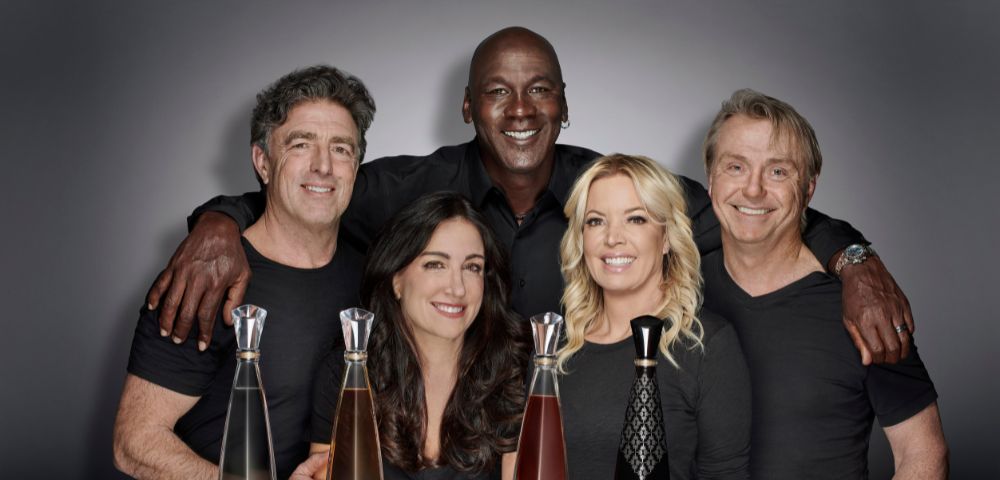 A diverse group of five people smiling warmly, posed behind four tall, elegant bottles of Cincoro Tequila. The setting is professional and inviting.
