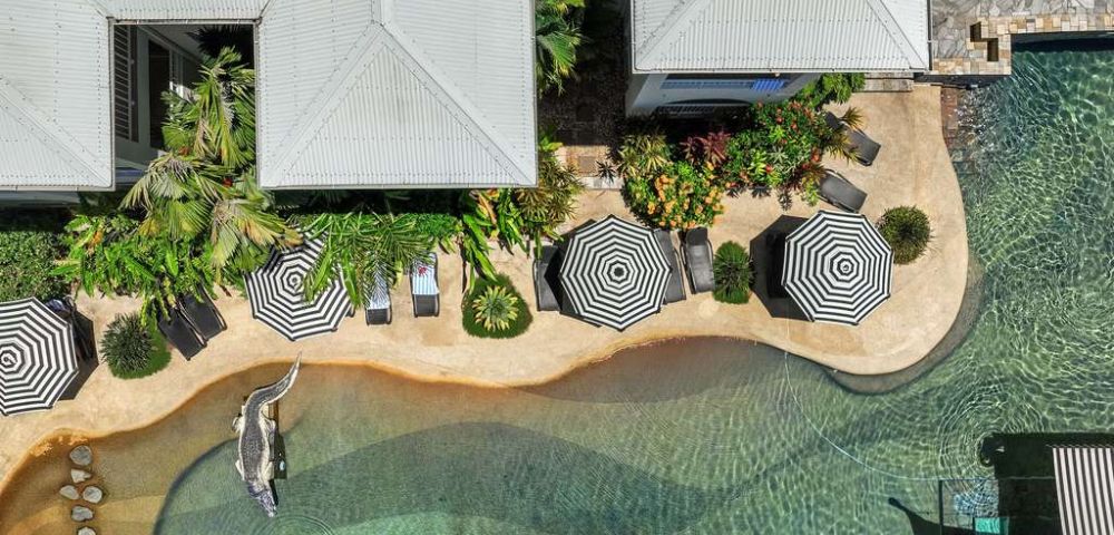 Aerial view of a resort with patterned umbrellas, a winding pool, lush greenery, and relaxed seating areas surrounding tranquil water.