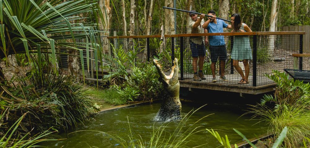 A crocodile leaps out of the water as three people watch from a deck in a lush, green enclosure surrounded by trees.