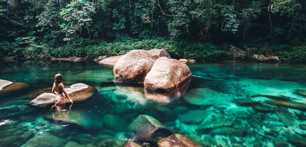 A girl stands on rocks in a clear turquoise river, surrounded by lush greenery and smooth river stones.