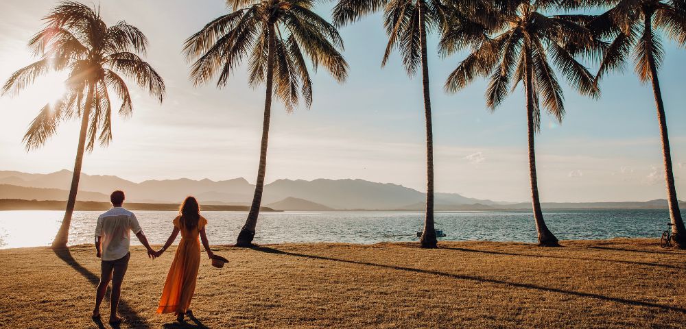 Couple holding hands, walking through palm trees at sunset, creating a romantic and serene atmosphere.
