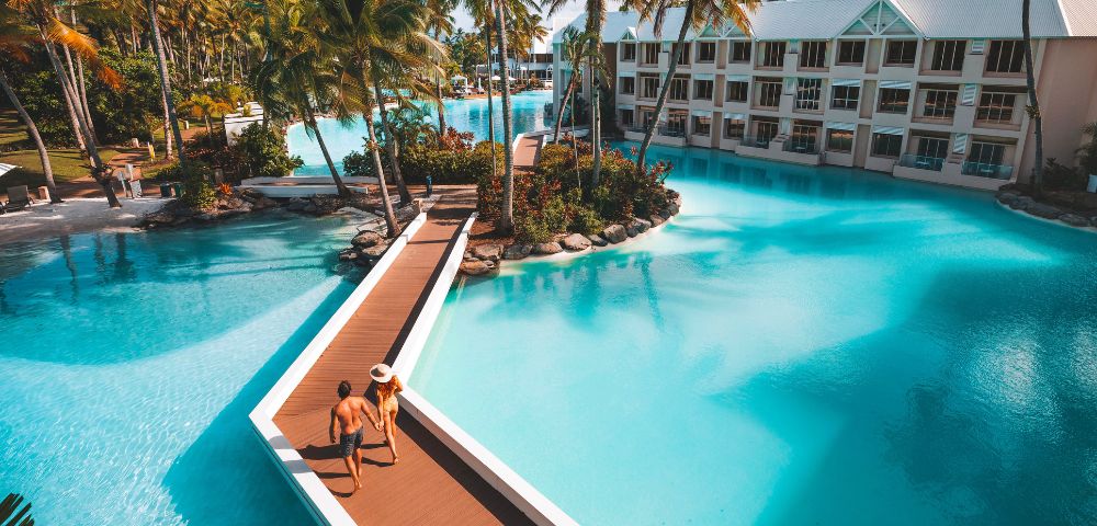 Tropical resort beach with soft sand, clear blue water, and palm trees under a bright sunny sky.