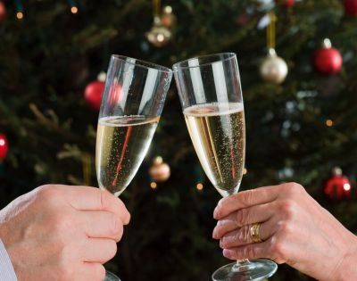 Two hands clink champagne glasses in a celebratory toast against a blurred background of a decorated Christmas tree with red and gold ornaments.