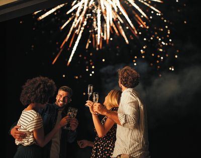 A group of four friends celebrate beneath a night sky lit by vibrant fireworks. They smile, holding champagne glasses, exuding joy and festivity.