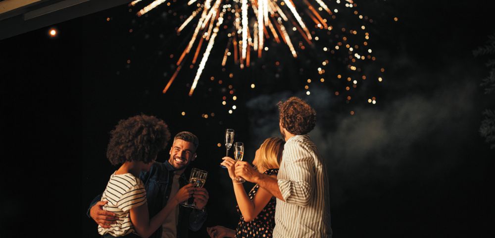 A group of four friends celebrate beneath a night sky lit by vibrant fireworks. They smile, holding champagne glasses, exuding joy and festivity.