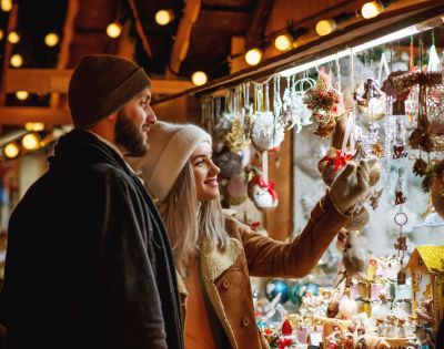 A couple in warm winter clothing enjoys browsing a festive market stall adorned with colorful holiday decorations, exuding a cozy and cheerful vibe.