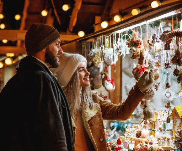 A couple in warm winter clothing enjoys browsing a festive market stall adorned with colorful holiday decorations, exuding a cozy and cheerful vibe.