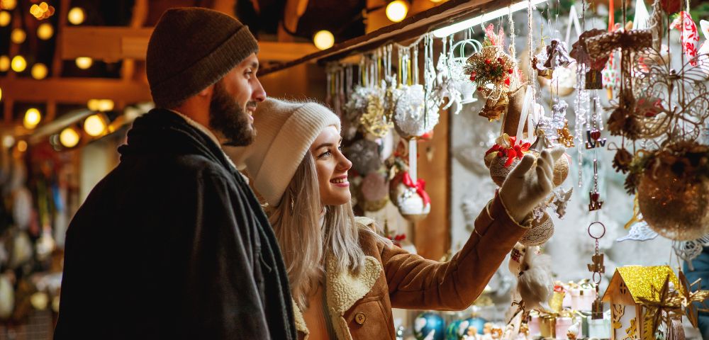 A couple in warm winter clothing enjoys browsing a festive market stall adorned with colorful holiday decorations, exuding a cozy and cheerful vibe.