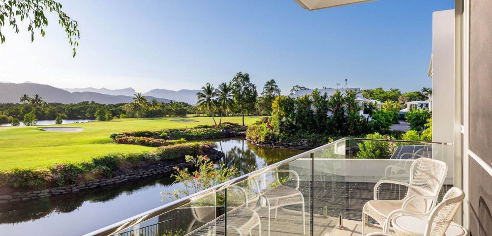 Balcony overlooking a serene golf course with lush greenery, a calm pond, and distant mountains under a clear blue sky, evoking tranquility.