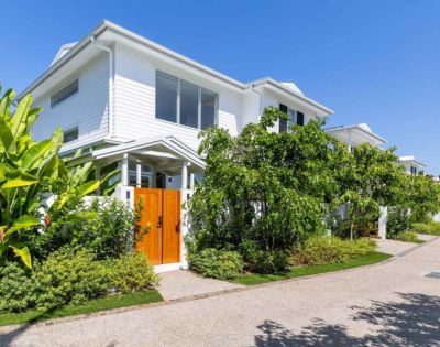 White two-story house with wooden doors, surrounded by lush green foliage under a clear blue sky. The scene is serene and tropical.