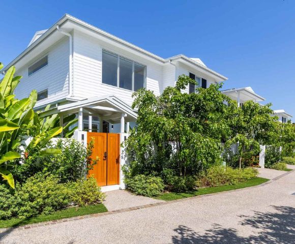 White two-story house with wooden doors, surrounded by lush green foliage under a clear blue sky. The scene is serene and tropical.