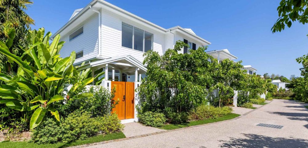 White two-story house with wooden doors, surrounded by lush green foliage under a clear blue sky. The scene is serene and tropical.
