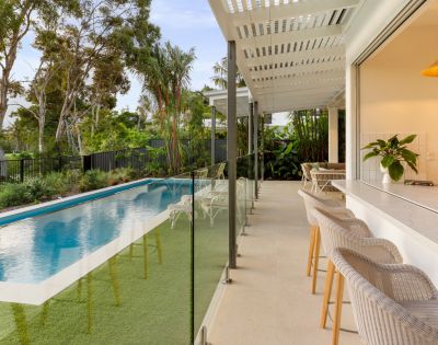 Modern patio with wicker bar stools beside a sleek swimming pool bordered by lush greenery. Large trees and clear blue sky set a serene tone.