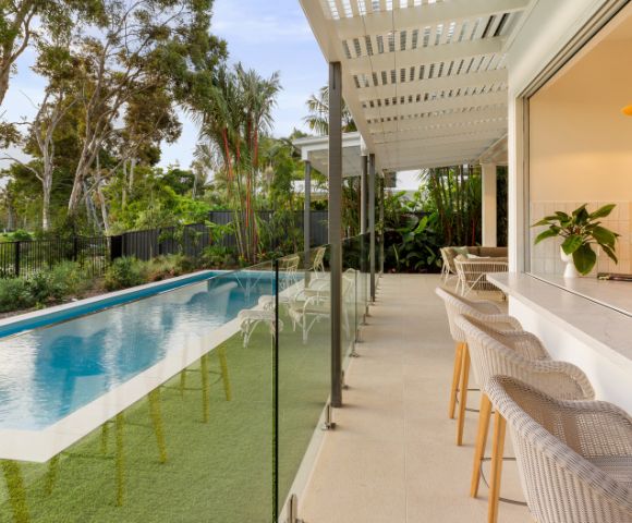 Modern patio with wicker bar stools beside a sleek swimming pool bordered by lush greenery. Large trees and clear blue sky set a serene tone.