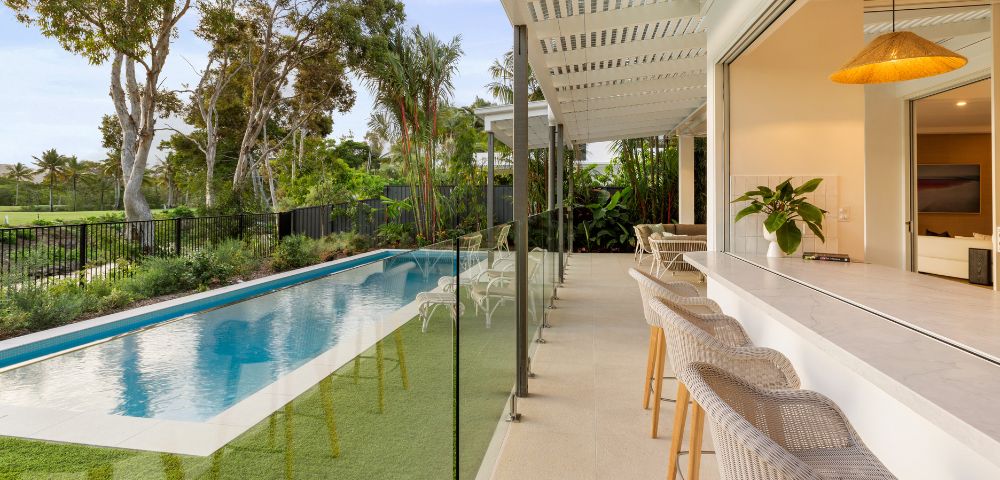 Modern patio with wicker bar stools beside a sleek swimming pool bordered by lush greenery. Large trees and clear blue sky set a serene tone.