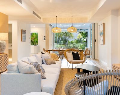 Bright, modern living room with neutral tones. Cozy white sofa, wicker chairs, wooden furniture, large windows revealing lush greenery outside. Warm and inviting.