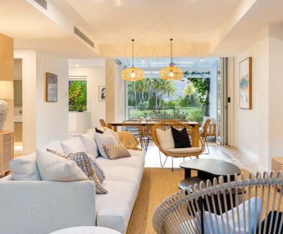 Bright, modern living room with neutral tones. Cozy white sofa, wicker chairs, wooden furniture, large windows revealing lush greenery outside. Warm and inviting.