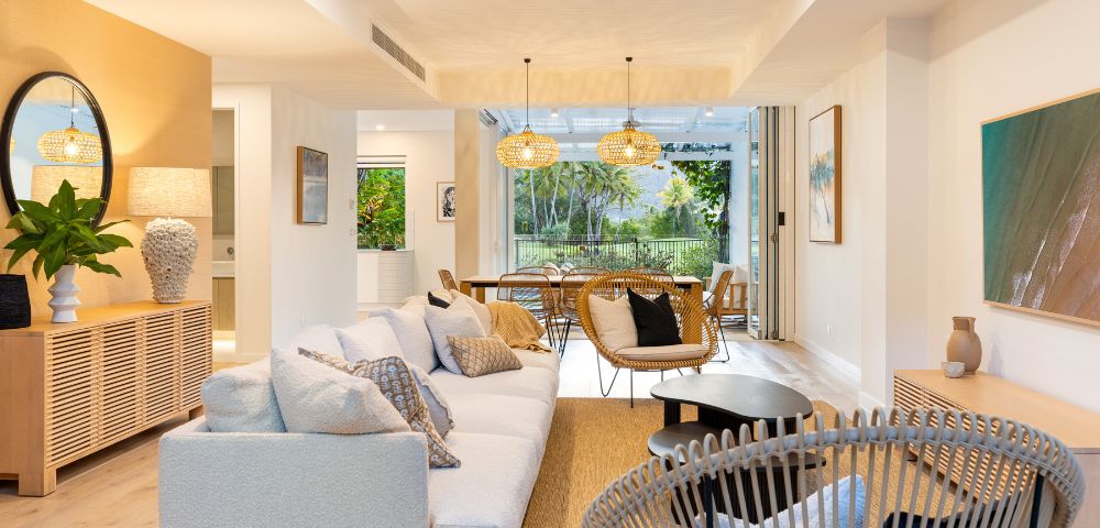 Bright, modern living room with neutral tones. Cozy white sofa, wicker chairs, wooden furniture, large windows revealing lush greenery outside. Warm and inviting.