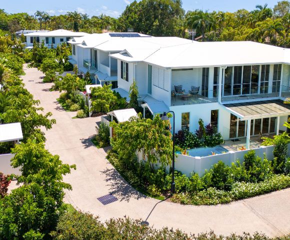 Modern tropical residential area, featuring white buildings with large glass windows, lush green gardens, and a clear blue sky, conveying tranquility.