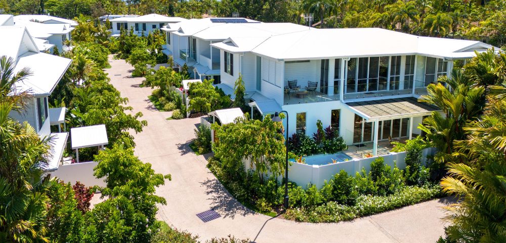 Modern tropical residential area, featuring white buildings with large glass windows, lush green gardens, and a clear blue sky, conveying tranquility.